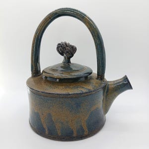 May include: A ceramic teapot with a dark blue and brown glaze. The teapot has a rounded body, a curved handle, and a spout. The lid is topped with a decorative knob. The overall design is rustic and handmade.
