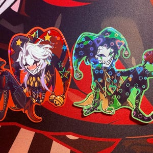 May include: Two colorful stickers featuring jester characters. One jester has white hair and a red and orange outfit, the other has dark hair and a green outfit. Both have wide smiles and star and heart details.