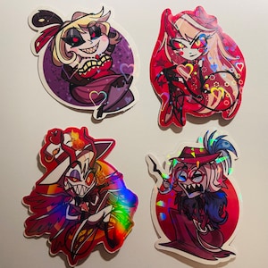 May include: Four holographic stickers featuring characters from the animated series Helluva Boss. The stickers are on a white background. The characters are:  a red-haired woman with horns, a woman with blonde hair and a red dress, a man with a red hat and a white shirt, and a man with a red hat and a black shirt.