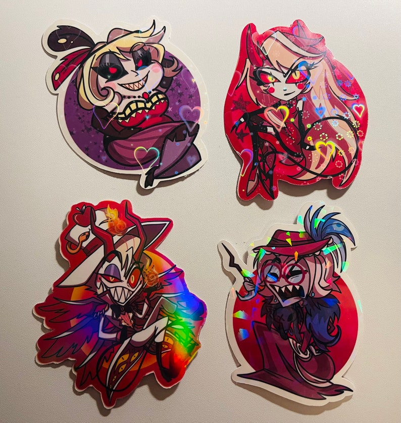 May include: Four holographic stickers featuring characters from the animated series Helluva Boss. The stickers are on a white background. The characters are:  a red-haired woman with horns, a woman with blonde hair and a red dress, a man with a red hat and a white shirt, and a man with a red hat and a black shirt.