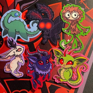 May include: A set of six colorful stickers featuring various creatures, including a green dinosaur, a black moth, a brown furry monster, a white rabbit, a purple demon, and a green monster with red eyes.