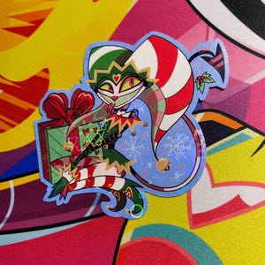 May include: A sticker featuring a cartoon jester character in a Christmas theme. The jester wears a green and red outfit, holding a wrapped gift. The background is light blue with snowflakes.