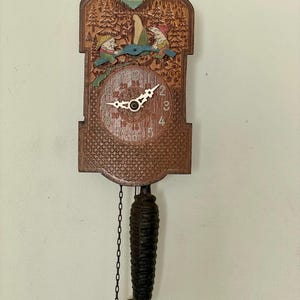 Puede incluir: Un reloj de cuco de madera vintage con un diseño tallado detallado. El reloj presenta una escena de bosque con figuras, una esfera circular y manecillas decorativas. Un péndulo en forma de piña cuelga debajo y el reloj está montado en una pared.