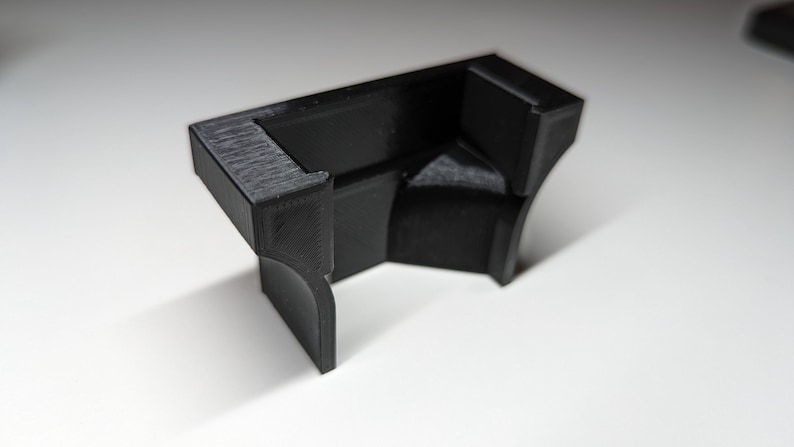 May include: A black, 3D-printed corner bracket with a textured surface. The bracket has a right-angle design with curved and angled edges. It is sitting on a white surface.