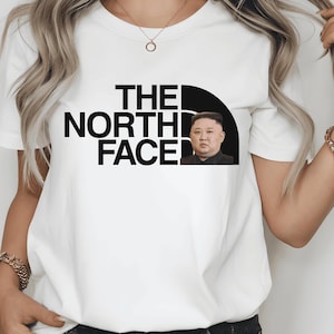 May include: White t-shirt featuring the text "THE NORTH FACE" in black, with a black and white graphic of a person's face. The shirt is a crew neck style.