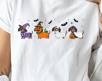 Op de afbeelding: Een wit T-shirt met korte mouwen en een afbeelding van vier teckels in Halloween-kostuums. De honden zijn verkleed als tovenaar, pompoen, mummie en spook, omringd door kleine zwarte vleermuizen en sterren op de voorzijde van het shirt.