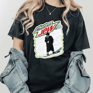 Puede incluir: Camiseta negra con una parodia del logo de Mountain Dew, con el texto "Mountain Jew". El gráfico muestra a un hombre con abrigo largo y sombrero sobre esquís en un paisaje nevado. La camiseta es de color oscuro.