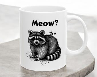 May include: White ceramic coffee mug featuring a black and white illustration of a raccoon and the text "Meow?" printed on it.