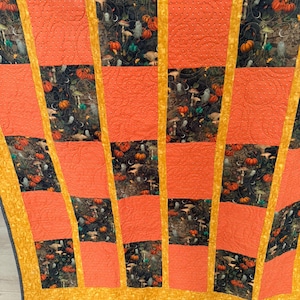 May include: A patchwork quilt featuring alternating rectangular panels of orange fabric with a subtle stippled texture and dark fabric printed with a pattern of pumpkins, mushrooms, and ghosts, all separated by thin yellow fabric borders.