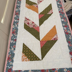 May include: A quilted table runner with a white center featuring a geometric design in shades of green, orange, and pink. The border is a floral pattern in blue, pink, and white, with a pink edge.