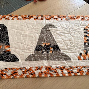 May include: A rectangular quilted table runner featuring three witch hat appliqués on a white background. The hats are patterned in black spiderwebs, gray moons, and orange plaid. The border displays a repeating print of white ghosts and bats.