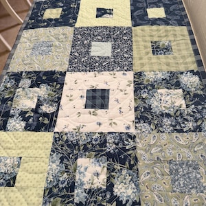 May include: A quilted table runner featuring a patchwork design. The runner showcases squares of various floral and paisley patterns in shades of blue, green, and white. The quilt is machine-stitched with a diamond pattern.