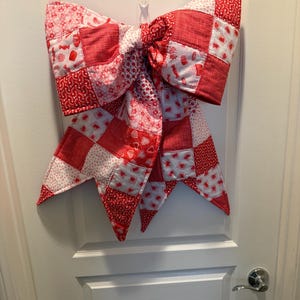 May include: A large, quilted bow in shades of red and white, hanging on a white door. The bow is made of patchwork squares with various patterns, including hearts and candy. The bow is a decorative item.