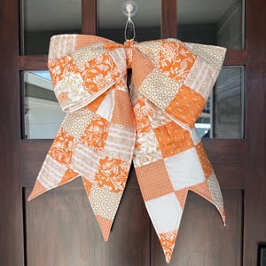 May include: A large decorative bow made of fabric with a patchwork design featuring various orange and white floral, geometric, and dotted patterns. The bow has two wide loops at the top and two long, pointed tails hanging down from the centre knot.