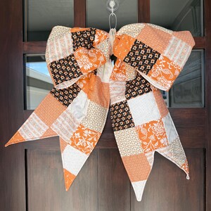 May include: A large decorative bow made of fabric with a patchwork design featuring various patterns in shades of orange, black, white, and cream. The bow has two wide loops and two long, pointed tails, all constructed from quilted squares of fabric.