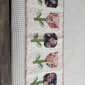 May include: A quilted table runner featuring a repeating floral tulip design. The tulips are made of fabric with floral patterns in shades of pink, blue, and red. The stems and leaves are green. The runner has a white background with a floral border.