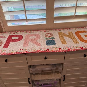 May include: A quilted table runner with the word "SPRING" in large, colorful letters. The letters are made of floral patterned fabric. A flower in a blue pot is between the letters "G" and "N". The runner has a floral border.