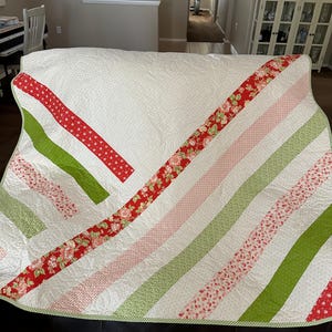 May include: A white quilt with diagonal stripes of red, green, and pink floral fabric. The quilt is folded and sitting on a brown leather couch.