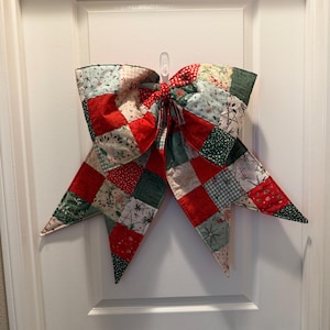 Puede incluir: Un gran lazo acolchado con un diseño de retazos, con telas rojas, verdes y azul claro. El lazo está colgado en una puerta blanca, creando una decoración festiva para las fiestas. El lazo mide aproximadamente 76 cm de ancho.