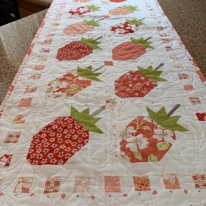 May include: A rectangular quilted table runner featuring a repeating pattern of strawberries in various shades of red, pink, and orange. Each strawberry has green fabric leaves and a brown stem, set against a white background with swirling stitching.