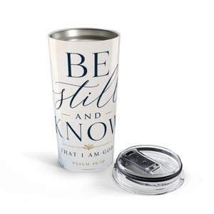 Op de afbeelding: Een witte roestvrijstalen beker met een donkerblauw en goudkleurig bedrukt ontwerp met de tekst BE STILL AND KNOW THAT I AM GOD PSALM 46:10. De beker heeft een taps toelopende cilindrische vorm en wordt geleverd met een doorzichtig plastic deksel.