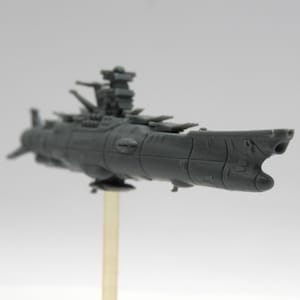 May include: A grey plastic model of a space battleship featuring a prominent bridge tower, multiple turret placements along the deck, and a pointed bow. The model is mounted on a thin, vertical support stand, showcasing its detailed hull structure.