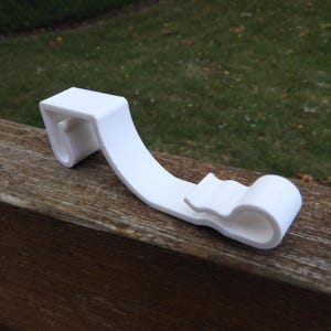 May include: White plastic hook designed to hang items on a fence or railing. The hook has a curved shape and a small lip to help secure items.