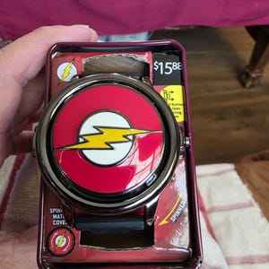 Puede incluir: Un reloj de pulsera redondo con correa negra y esfera circular roja con un emblema de rayo amarillo. El reloj se presenta en una caja de cartón granate con texto que incluye $15.88, Spinner y un icono de un rayo.