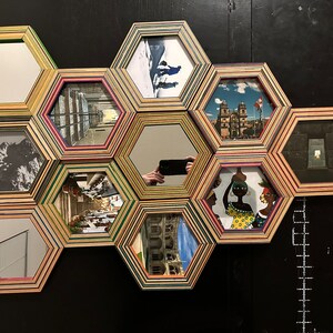 Pu&ograve; includere: Un'opera d'arte murale composta da cornici esagonali, ciascuna con un'immagine o uno specchio diverso. Le cornici sono in legno e presentano un design multistrato colorato. Le immagini includono una scena di montagna innevata, una strada cittadina, una chiesa, una persona che tiene una brocca e uno specchio che riflette una persona che scatta una foto.