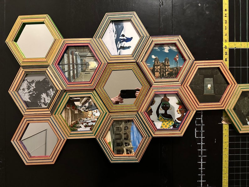 Pu&ograve; includere: Un'opera d'arte murale composta da cornici esagonali, ciascuna con un'immagine o uno specchio diverso. Le cornici sono in legno e presentano un design multistrato colorato. Le immagini includono una scena di montagna innevata, una strada cittadina, una chiesa, una persona che tiene una brocca e uno specchio che riflette una persona che scatta una foto.
