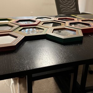 Pu&ograve; includere: Un set di 12 cornici per foto esagonali in legno con un design a strisce multicolori. Le cornici sono disposte a nido d'ape su un tavolo nero.