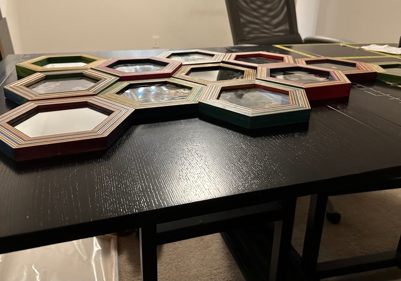Pu&ograve; includere: Un set di 12 cornici per foto esagonali in legno con un design a strisce multicolori. Le cornici sono disposte a nido d'ape su un tavolo nero.