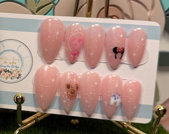 May include: A set of ten almond-shaped press-on nails in a light pink shade with white polka dots, featuring small character decals. The nails are displayed on a rectangular card next to a white ornate picture frame containing a floral logo.