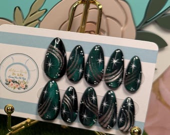 Op de afbeelding: Een set van tien amandelvormige press-on nagels met een donkergroen en zwart verloop, zilveren wervelende lijnen en witte steraccenten, weergegeven op een witte kaart naast een witte, sierlijke fotolijst met een bloemenlogo.