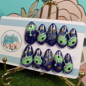 May include: A set of ten almond-shaped press-on nails featuring a dark blue starry background with green three-eyed alien character graphics. The nails are displayed on a white card held by a gold stand next to a white ornate picture frame.