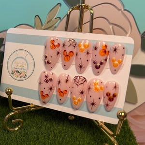 May include: A set of ten almond-shaped press-on nails featuring a light pink base with orange pumpkin-shaped icons, black spiderweb designs, and black starburst accents. The nails are displayed on a white card held by a gold metal stand.