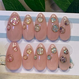 May include: A set of ten almond-shaped press-on nails in a soft pink hue, each decorated with small, iridescent circular accents. Beside the nails sits a white ornate picture frame containing a card with the text Artisan Nails By Julia.