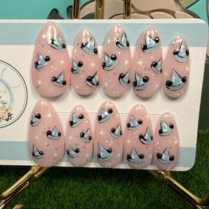 May include: A set of ten almond-shaped press-on nails with a light pink base, featuring a repeating pattern of small blue witch hats and white starburst accents. The nails are displayed on a white card with the text Artisan Nails By Julia.