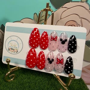 May include: A set of ten almond-shaped press-on nails displayed on a white card. The nails feature red with white polka dots, light pink with black Mickey Mouse silhouettes, light pink with red bows, and black with white polka dots designs.