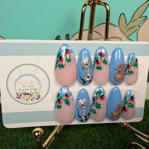 Puede incluir: Un juego de diez uñas postizas en forma de almendra con base azul claro y nude. Los diseños incluyen ratones grises pintados a mano con gorros de chef blancos, hojas de acebo verdes y pequeñas bayas rojas. Las uñas se presentan en una tarjeta.