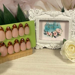 May include: Artificial nails with a pale pink base and a brown leopard print tip design. The nails are displayed on a green card. A white ornate frame with the text "Artisan Nails" is in the background.
