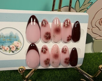 May include: A set of ten almond-shaped press-on nails featuring a light pink base. The design includes deep burgundy French tips, solid burgundy nails, and light pink nails decorated with burgundy floral patterns and gold starburst accents.