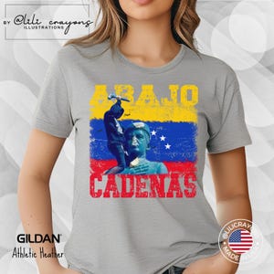 May include: A gray t-shirt with a graphic design featuring a person breaking chains with a hammer. The graphic includes the text "ABAJO CADENAS" and the colors of the Venezuelan flag.