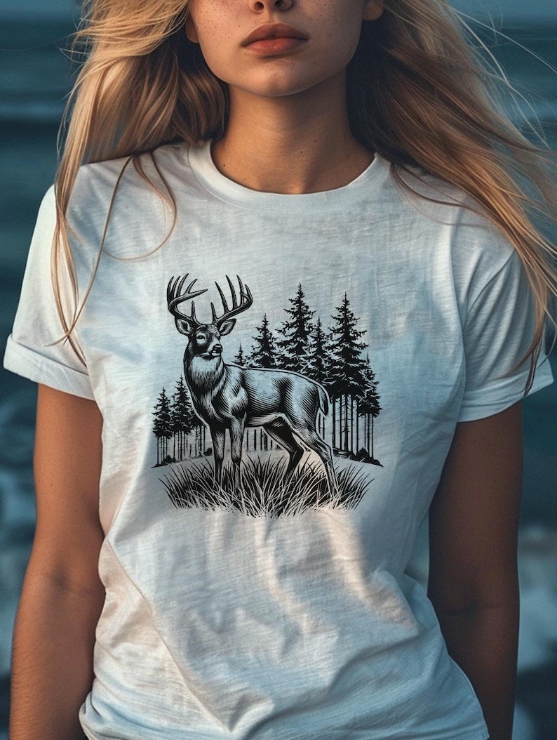 Może przedstawiać: Biały t-shirt z czarno-białą ilustracją jelenia stojącego w lesie. Jeleń ma duże poroże i jest otoczony sosnami.