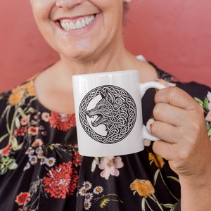 May include: A white ceramic mug with a black and white design of a wolf's head in a Celtic knot. The mug is being held by a person wearing a floral dress.