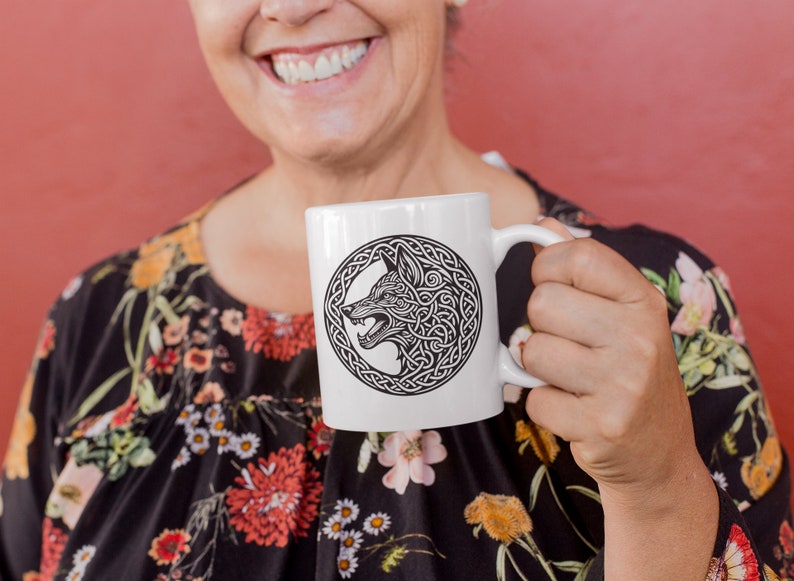 May include: A white ceramic mug with a black and white design of a wolf's head in a Celtic knot. The mug is being held by a person wearing a floral dress.