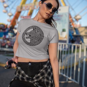 May include: A woman wearing a gray cropped t-shirt with a black and white wolf design in a Celtic knot pattern. She is wearing black shorts and a plaid shirt tied around her waist. She is standing in front of a blurry background of a Ferris wheel.