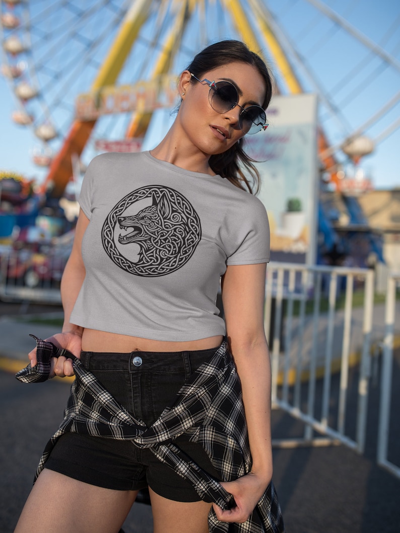 May include: A woman wearing a gray cropped t-shirt with a black and white wolf design in a Celtic knot pattern. She is wearing black shorts and a plaid shirt tied around her waist. She is standing in front of a blurry background of a Ferris wheel.