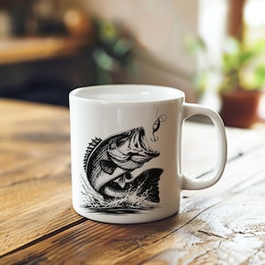 K&ouml;nnte beinhalten: Wei&szlig;e Keramiktasse mit einer Schwarzwei&szlig;illustration eines Barsches, der mit einem K&ouml;der im Maul aus dem Wasser springt.