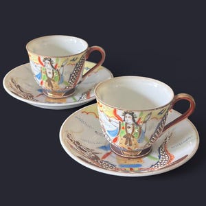 May include: Two porcelain teacups and saucers. The cups have a white interior and are decorated with a colorful illustration of a figure. The saucers feature a dragon design. The cups have brown handles and are set against a dark background.
